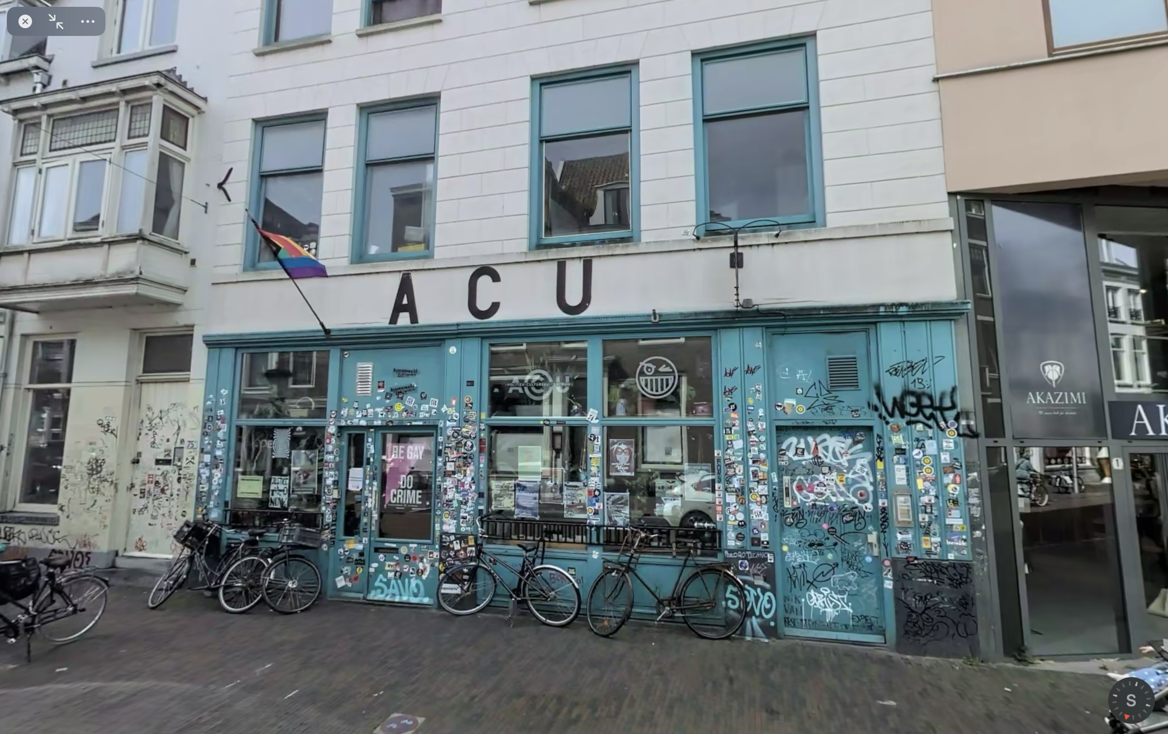 Photo of depicting the entrance of ACU in Utrecht: a ground-floor bar with many leftist stickers on the windows, and a queer flag above, with a standard second and third floor wall and windows above